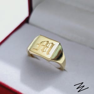 May include: A gold signet ring with the letter "M" engraved in an elegant, stylized font. The ring has a rectangular face and a polished finish, presented in a red and white ring box.