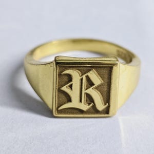 May include: A gold-toned signet ring featuring a square face with a textured background. The center of the ring displays a prominent, raised letter R in an old English gothic font style. The band is smooth and tapers toward the back of the ring.