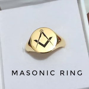 May include: A gold-colored Masonic ring with a round face featuring a black square and compass symbol. The ring is presented in a white box, with the words "MASONIC RING" printed below.