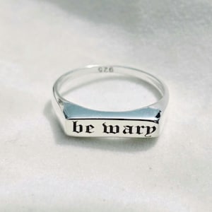 May include: A polished silver ring featuring a flat rectangular face engraved with the black text be wary in a gothic font. The interior of the band is clearly stamped with the number 925, indicating sterling silver composition for this piece.
