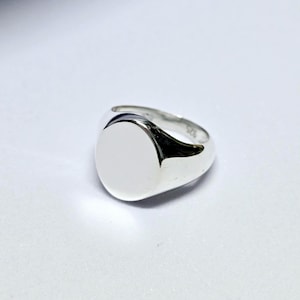May include: A polished silver signet ring with a smooth, oval face. The ring has a rounded band and a bright, reflective surface. The design is simple and classic, suitable for everyday wear. The ring is a size 9.