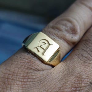 Könnte beinhalten: Ein goldener Siegelring mit einer quadratischen Fläche, auf der ein gravierter Buchstabe "P" zu sehen ist.