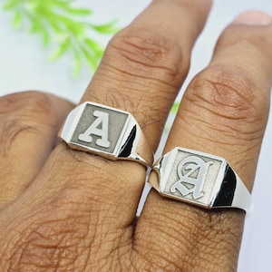 May include: Two silver signet rings, each with the letter "A". One ring has a block letter "A" and the other has a stylized "A". The rings are on a person's fingers.