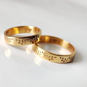 May include: Two gold wedding rings with leaf patterns and engraved initials and a heart symbol. The rings are displayed on a white surface, reflecting light. The bands are a warm, shiny gold colour.