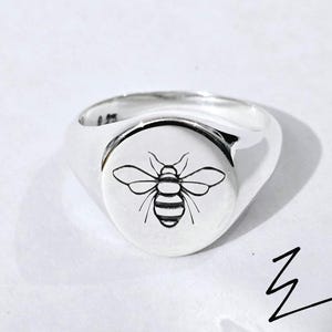 Könnte beinhalten: Ein silberner Siegelring mit rundem Gesicht, der eine schwarz-weiße Bienenillustration zeigt. Der Ring hat eine polierte Oberfläche und ein breites Band. Das Bienendesign ist auf der Vorderseite des Rings zentriert.