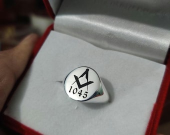 May include: A silver ring with a round face featuring a black Masonic symbol and the number 1045. The ring is presented in a red jewelry box with a white satin interior.
