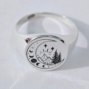 May include: A silver signet ring with a detailed black and white design. The design features a crescent moon, mountains, stars, and trees. The ring has a smooth, polished finish.