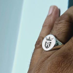 May include: A silver ring with an oval-shaped top featuring a shield with the letter "R" and a crown above it.