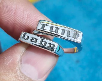 May include: Two silver-toned metal rings with flat rectangular faces. The top ring is engraved with the word "cunt" in a black gothic-style font, and the bottom ring is engraved with the word "baby" in the same black gothic-style font.