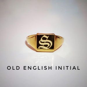 May include: A gold signet ring with a square face featuring the Old English letter "S" in black. The ring has a polished finish and is set against a white background. The text "OLD ENGLISH INITIAL" is below the ring.