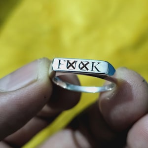 May include: A silver ring with the word "FXXK" engraved in black, held between fingers. The ring has a rectangular top and a polished finish, set against a yellow background.