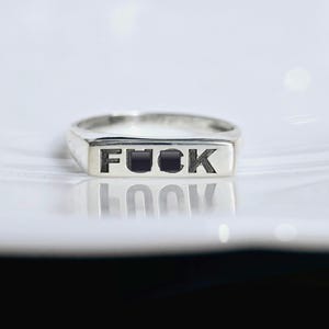 May include: A silver ring with the word "FUCK" engraved in black block letters. The ring has a rectangular face and a polished finish, reflecting the light. The ring is displayed on a white surface.
