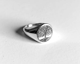 May include: A silver signet ring with a detailed tree of life design. The ring has a polished finish and a rounded, oval face with the tree's branches and roots intricately carved.