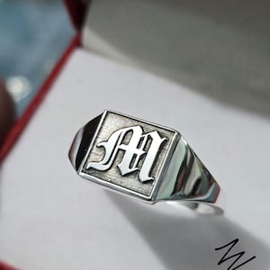 May include: A silver signet ring with a square face featuring the capital letter "M" in a gothic font. The ring is set in a red box, showcasing its polished, reflective surface. The ring is a classic piece of jewelry.