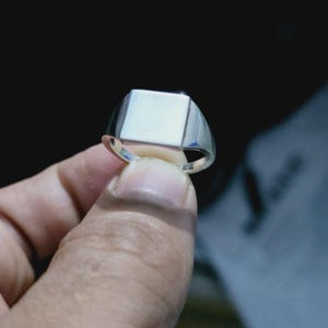 May include: A polished silver-tone signet ring featuring a smooth, flat, rectangular face and a solid, tapered band. The metal surface has a reflective, metallic finish that catches the light, showcasing a clean, minimalist design with no engravings.