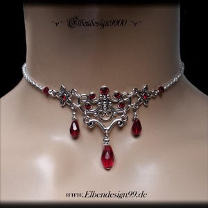 Puede incluir: Collar gargantilla plateado con detalles de gemas rojas. El collar presenta una pieza central decorativa con gemas colgantes en forma de lágrima. El diseño incluye detalles florales y de volutas, con el texto "Elbendesign9900" y "www.Elbendesign99.de".