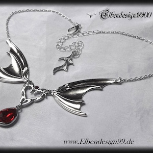 May include: Silver necklace featuring two bat wings and a dangling red gemstone. The necklace has a delicate chain and a small bat wing charm.