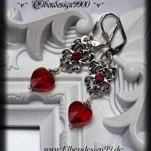 Peut inclure: Boucles d'oreilles pendantes argentées avec un motif filigrané, ornées d'une petite pierre rouge et d'un pendentif en cristal rouge en forme de cœur. Les boucles d'oreilles sont présentées sur un cadre décoratif blanc. Le site web www.Elbendesign99.de est visible.