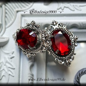 May include: Two silver-coloured brooches with large, oval, deep red gemstones. The brooches feature intricate filigree detailing around the gems and are connected. The image includes the text "Elbendesign9900" and "www.Elbendesign99.de".