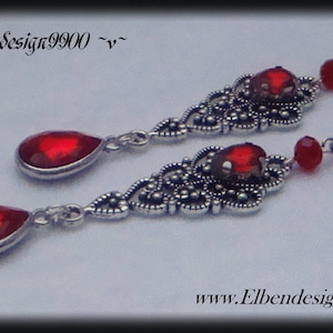 May include: A pair of silver dangle earrings featuring red teardrop-shaped crystals. The earrings have a filigree design and are accented with smaller red beads.