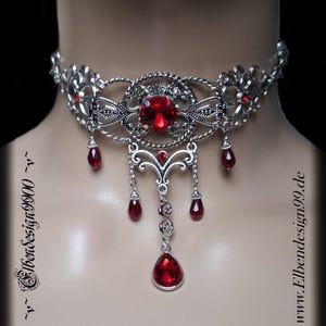Può includere: Collana girocollo argentata con una grande gemma rossa rotonda al centro e gemme rosse più piccole a forma di goccia. L'intricato design a filigrana presenta un ciondolo centrale e accenti pendenti.
