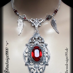 May include: Silver necklace featuring a large, oval, red gemstone pendant with an ornate filigree design. The necklace has two small wing charms and red bead accents. The website address "Elbendesign99.de" is visible.