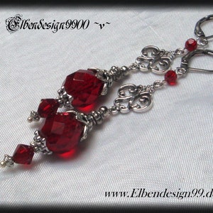 Può includere: Orecchini d'argento con perline di vetro rosso e accenti argentati decorativi. Gli orecchini presentano un design pendente con più perline e un ciondolo d'argento decorativo.
