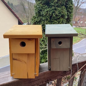 May include: Two wooden birdhouses, one is natural wood colour and the other is painted a dark green. Both birdhouses have a small round opening for birds to enter.