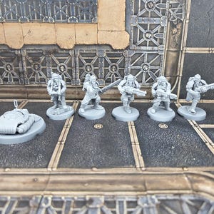 Op de afbeelding: Een verzameling ongeverfde, grijze miniatuurfiguren, waaronder soldaten met geweren en een klein voertuig. De figuren staan op individuele bases, wat suggereert dat ze bedoeld zijn voor tabletop gaming of modelbouw. De scène speelt zich af op een gedetailleerd spelbord met industriethema.