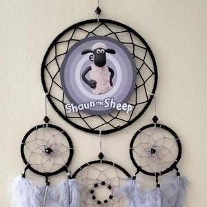 以下が含まれることがあります： 黒いウェブデザインとグレーと白の羽が特徴の手作りのドリームキャッチャー。 上部の円にはショーン・ザ・シープのグラフィックがあります。 「TOM」という名前が中央の円に白い文字で表示されています。