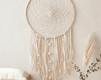 Puede incluir: Atrapasueños color crema con un diseño de telaraña circular tejido. El atrapasueños presenta largas cintas fluidas y pequeños adornos redondos colgando de la parte inferior. Se suspende contra una pared de tonos neutros, creando una estética bohemia.