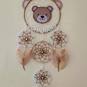 Puede incluir: Un atrapasueños marrón y blanco con una cara de oso en el centro. El atrapasueños tiene cuatro círculos más pequeños con una red marrón y blanca y plumas que cuelgan de la parte inferior. El atrapasueños tiene el nombre "JOHN HANS" escrito en él.