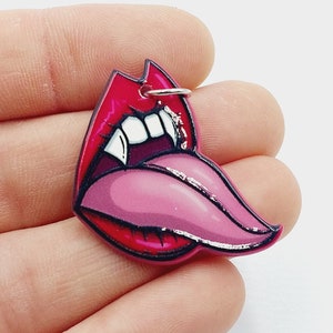 May include: A red and pink acrylic charm featuring a cartoon image of a mouth with fangs and a tongue sticking out. The charm has a silver ring for attaching to a keychain or other items.