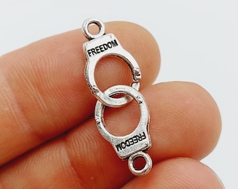 Pode incluir: Encanto de metal prateado em forma de algemas com a palavra "FREEDOM" gravada em cada lado.