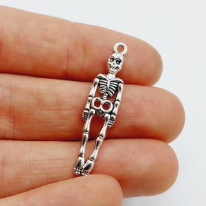 May include: Silver skeleton charm pendant. The skeleton is standing with its arms at its sides. The charm is about 2 inches tall.