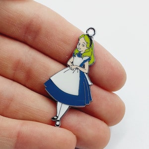 May include: A metal charm depicting Alice from Alice in Wonderland. Alice is shown in her blue dress with a white apron and blonde hair. The charm has a silver loop at the top for attaching to jewellery or crafts. The charm is a colourful and detailed representation.