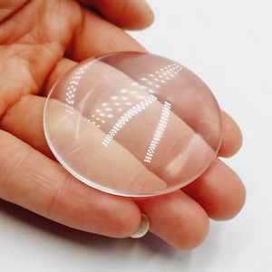 Op de afbeelding: Een heldere, cirkelvormige lens met een licht convexe oppervlakte. De lens wordt in een hand gehouden tegen een witte achtergrond. De lens lijkt de details erin te vergroten. De afbeelding is goed verlicht, wat de helderheid van het glas benadrukt.