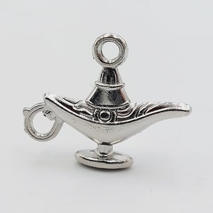 Puede incluir: Un amuleto de plata con forma de lámpara de genio. La lámpara tiene una parte superior redonda con un bucle para sujetarlo a una pulsera o collar. La lámpara tiene detalles intrincados y un pico curvo.
