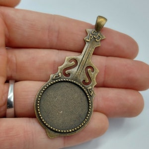May include: A bronze-colored pendant shaped like a violin with a round, blank cameo. The cameo is surrounded by a beaded border. The violin has musical notes engraved on the neck.