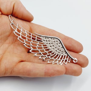 Op de afbeelding: Zilveren metalen vleugelbedel met een gedetailleerd, ingewikkeld ontwerp. De vleugel heeft een gaasachtig patroon en een kleine lus aan het einde.