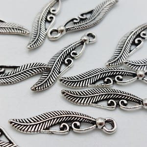 Op de afbeelding: Een groep zilverkleurige metalen bladcharms met ingewikkelde ontwerpen. De charms hebben een lus aan de bovenkant om aan sieraden of andere knutselwerken te bevestigen.