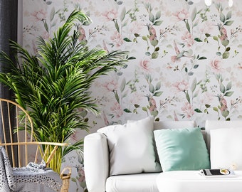 Op de afbeelding: Een witte bank met mintgroene en oudroze kussens, een witte salontafel met een roze bolvormige voet, een wit kleed, een houten stoel met een grijze plaid en behang met een herhalend patroon van roze bloemen en groene bladeren.