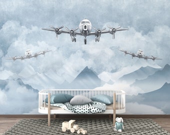 May include: A wall mural featuring three vintage-style propeller aeroplanes flying over a mountain range and clouds. The scene is rendered in shades of blue and white. Below the mural sits a white wooden bed frame with pillows and a blanket.