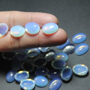 May include: A collection of oval-shaped, iridescent opal cabochons. The cabochons are a pale blue color with a white opalescence. The cabochons are arranged in a pile on a dark surface.