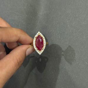Könnte beinhalten: Ein goldener Ring mit einem marquiseförmigen, tiefroten Edelstein, umgeben von einem Kranz aus kleinen, klaren Steinen. Der Ring hat ein schlichtes, poliertes Goldband. Die Fassung ist ein klassisches Design, geeignet für verschiedene Anlässe.