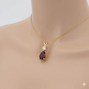 Puede incluir: Un collar con colgante de oro con una piedra preciosa roja oscura en forma de pera debajo de una piedra preciosa redonda y transparente. El colgante cuelga de una fina cadena de oro. El collar se muestra sobre un fondo neutro.