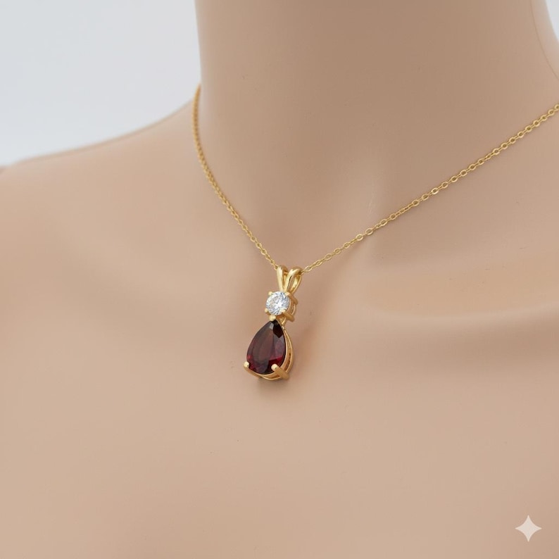 Puede incluir: Un collar con colgante de oro con una piedra preciosa roja oscura en forma de pera debajo de una piedra preciosa redonda y transparente. El colgante cuelga de una fina cadena de oro. El collar se muestra sobre un fondo neutro.