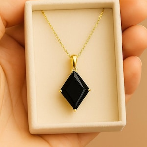 Puede incluir: Un collar dorado con un colgante negro en forma de diamante. El colgante tiene detalles dorados en cada esquina. El collar se presenta en una caja de color crema.