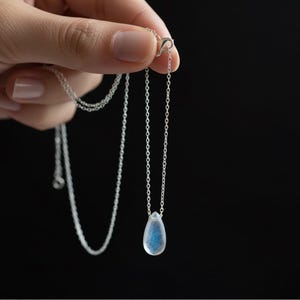May include: A silver necklace featuring a teardrop-shaped, iridescent blue gemstone pendant. The necklace is displayed against a black backdrop, highlighting the pendant's subtle colour variations and the chain's fine links. Jewellery.
