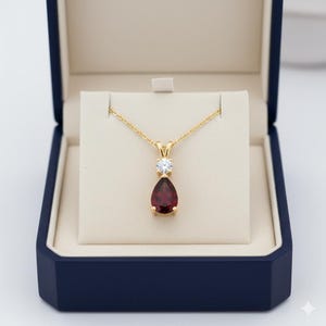 Puede incluir: Un collar dorado con un colgante de piedra preciosa roja oscura en forma de pera y una pequeña piedra preciosa clara y redonda. El collar se exhibe en una caja de joyería azul con interior crema.